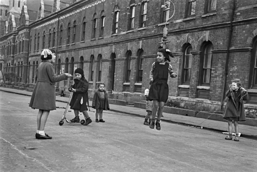 Six children play in the street outside a row of brick buildings; two girls jump rope, others watch or play, one uses a scooter. It’s a lively, vintage city scene.