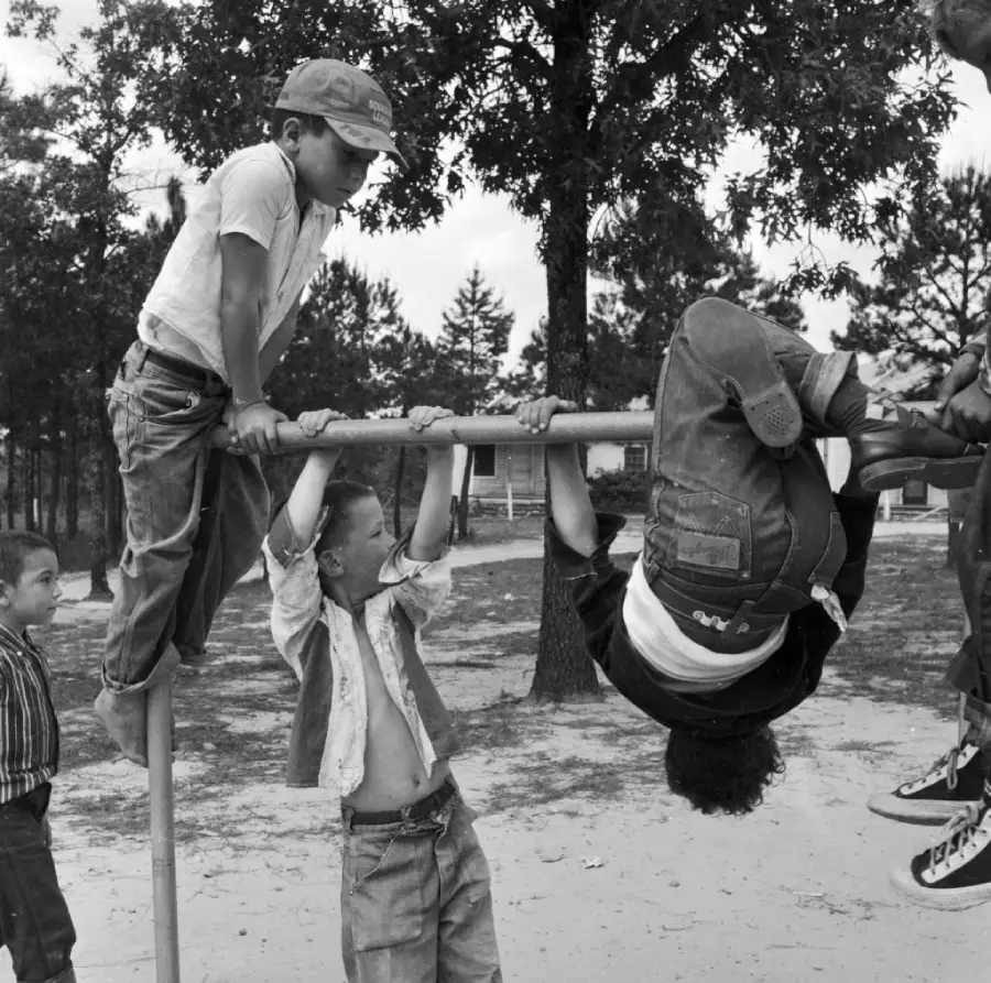 Three children play on a metal bar outdoors, with one hanging upside down and two others climbing or holding on, while another child watches nearby. Trees and a building are visible in the background.