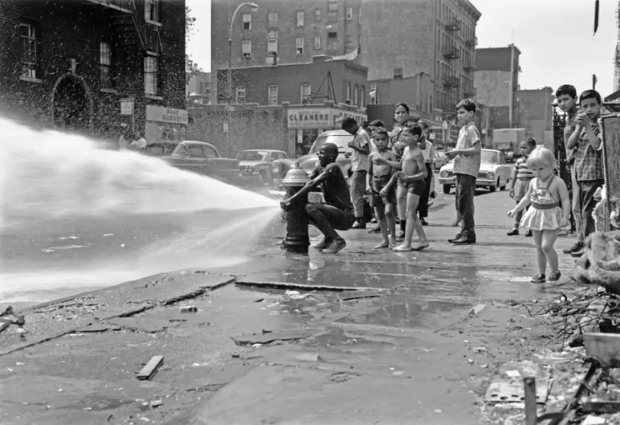 Children play in the spray of an open fire hydrant on a city street, while adults and kids watch. Buildings, cars, and debris are visible in the background.