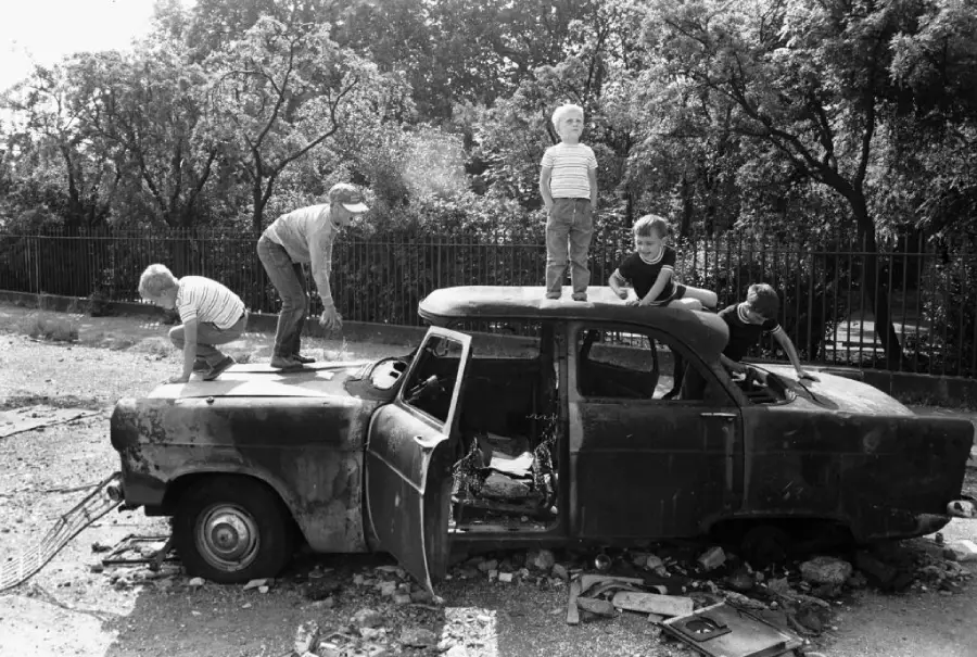 Five children play on and inside an old, rusted, abandoned car in an outdoor area with trees and a metal fence in the background. Sunlight casts shadows on the ground.