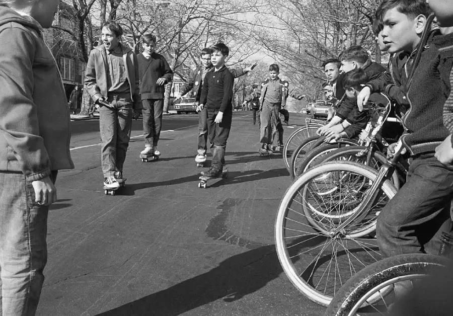A group of boys ride skateboards and bicycles on a suburban street lined with bare trees and parked cars, enjoying a sunny day.