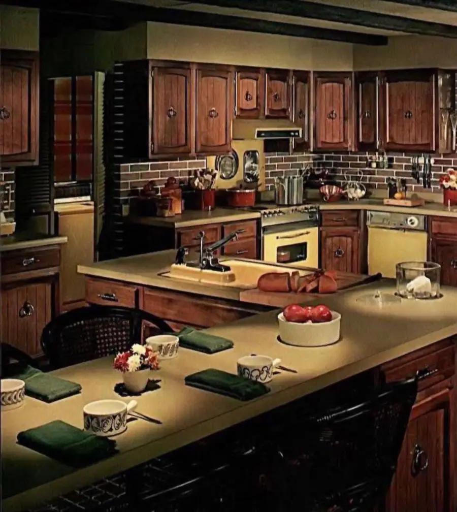 A vintage kitchen with wood cabinets, brown countertops, retro appliances, brick-patterned backsplash, and an island set with mugs, bowls, napkins, and a breadboard. Warm lighting gives a cozy atmosphere.