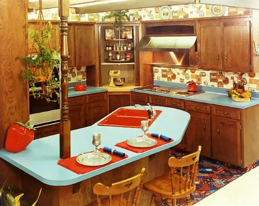 A retro kitchen with wood cabinets, blue countertops, red sink, patterned wallpaper, and a breakfast bar set for two. Hanging plants, cookbooks, and vintage decor complete the cozy, colorful space.
