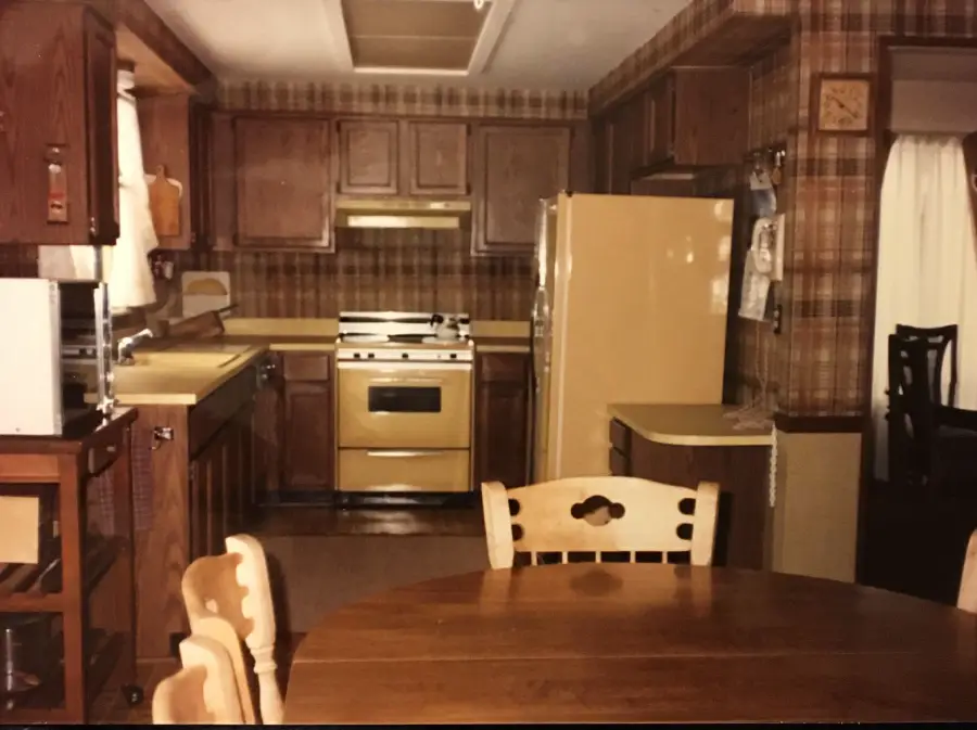 A vintage kitchen with brown wood cabinets, plaid wallpaper, yellow appliances, and a round wooden dining table with chairs in the foreground.