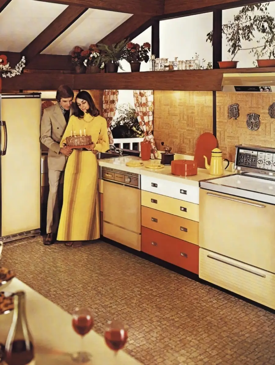 A couple stands in a retro 1970s kitchen, holding a birthday cake with candles. The kitchen features yellow, orange, and cream appliances, wood accents, and potted plants.