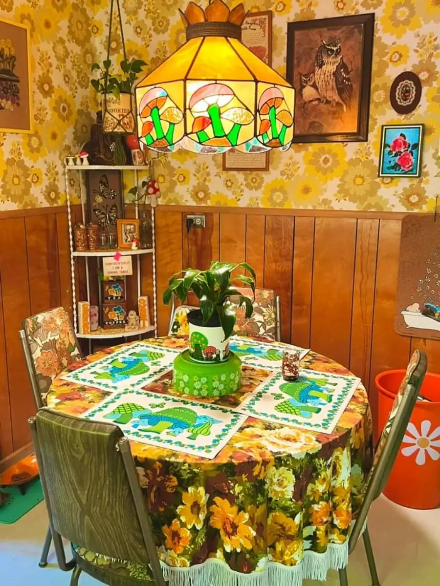 A retro dining nook with wood paneling, floral wallpaper, a round table with a colorful tablecloth, green chairs, a stained-glass lamp, houseplants, and eclectic 1970s-style decor on the walls and shelves.