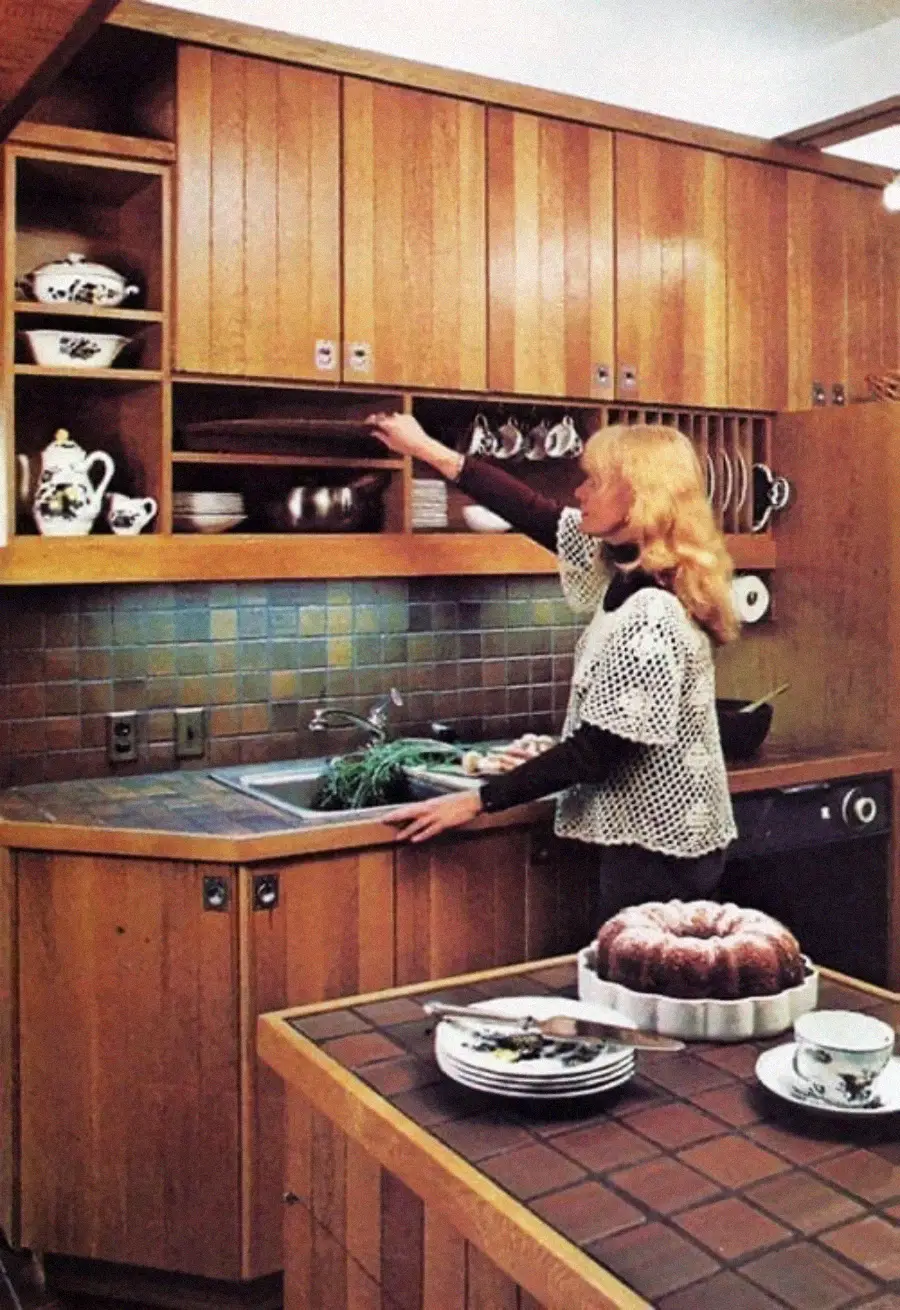 A woman in a vintage kitchen reaches for a shelf above a tiled countertop, with a bundt cake, plates, cups, and a teapot arranged on the counter in the foreground.