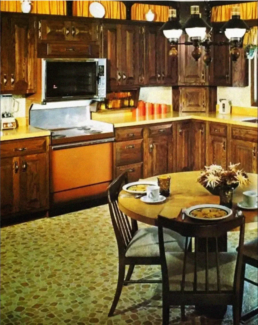 A vintage kitchen with dark wood cabinets, orange appliances, a round table with four chairs, dishes, and flowers. The floor has a patterned linoleum design, and a retro light fixture hangs overhead.