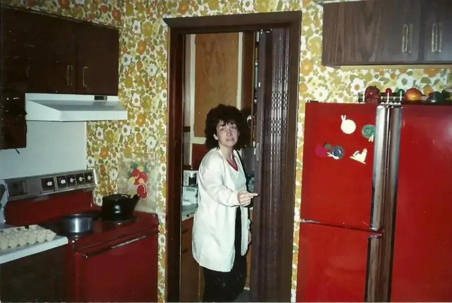 A woman stands in a retro kitchen with floral wallpaper, red cabinets, and a red refrigerator decorated with magnets. She wears glasses and a white jacket, looking toward the camera while holding the doorframe.