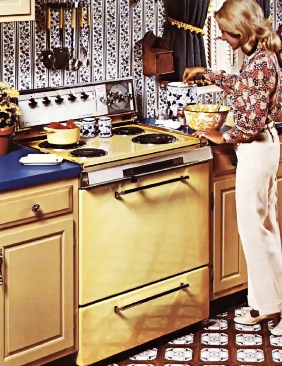 A woman prepares food in a vintage kitchen with a yellow stove, blue counter, floral wallpaper, and patterned floor tiles.