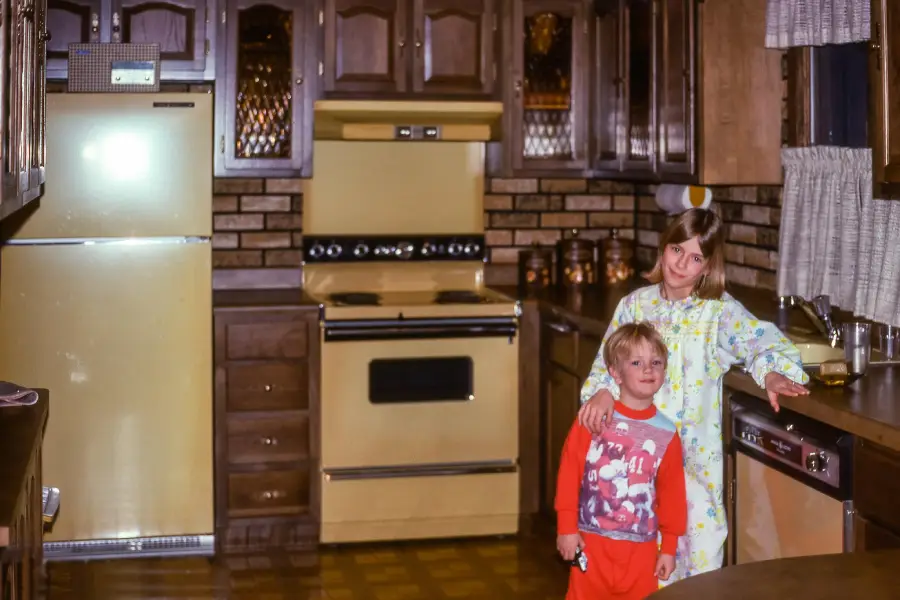 Two children in pajamas stand in a vintage kitchen with brown cabinets, a yellow fridge, and a matching stove. The girl hugs the younger boy, and both look at the camera. The kitchen has brick backsplash and patterned linoleum flooring.