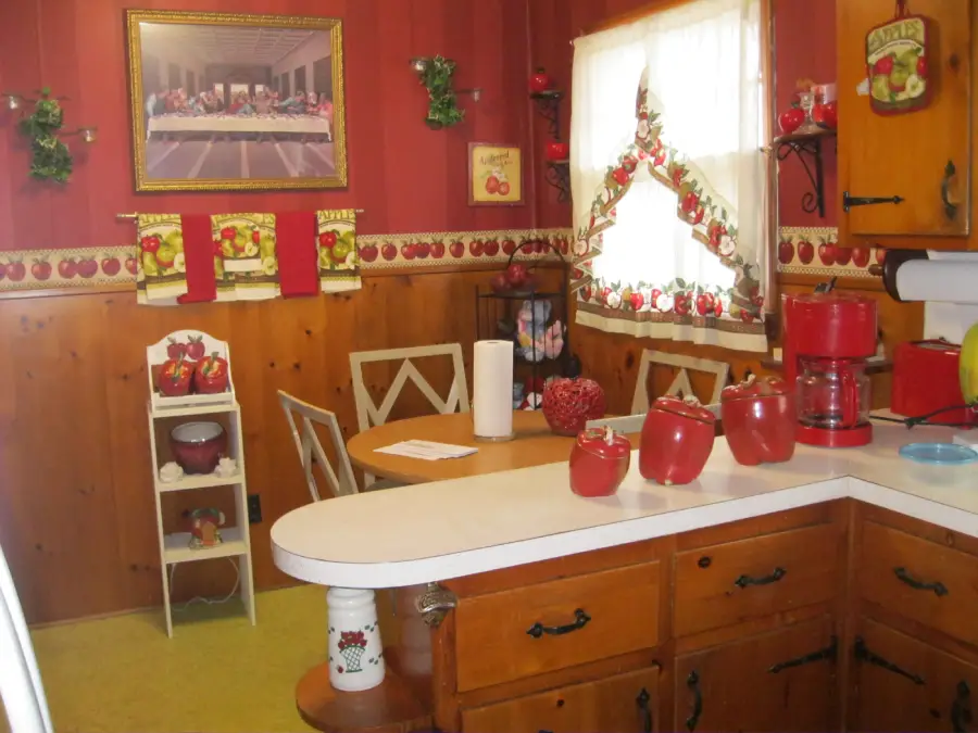 A cozy kitchen with red and wooden decor, apple-themed containers, a coffee maker, and curtains. A framed picture hangs above a table and chairs near a window with bright light.