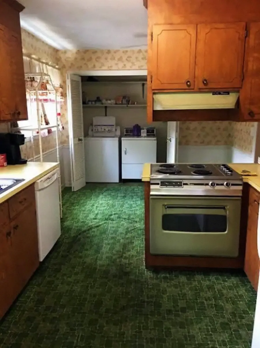 Retro kitchen with green carpet, wood cabinets, and an olive green stove. A washing machine and dryer are visible in an adjacent laundry area. Light wallpaper decorates the walls.