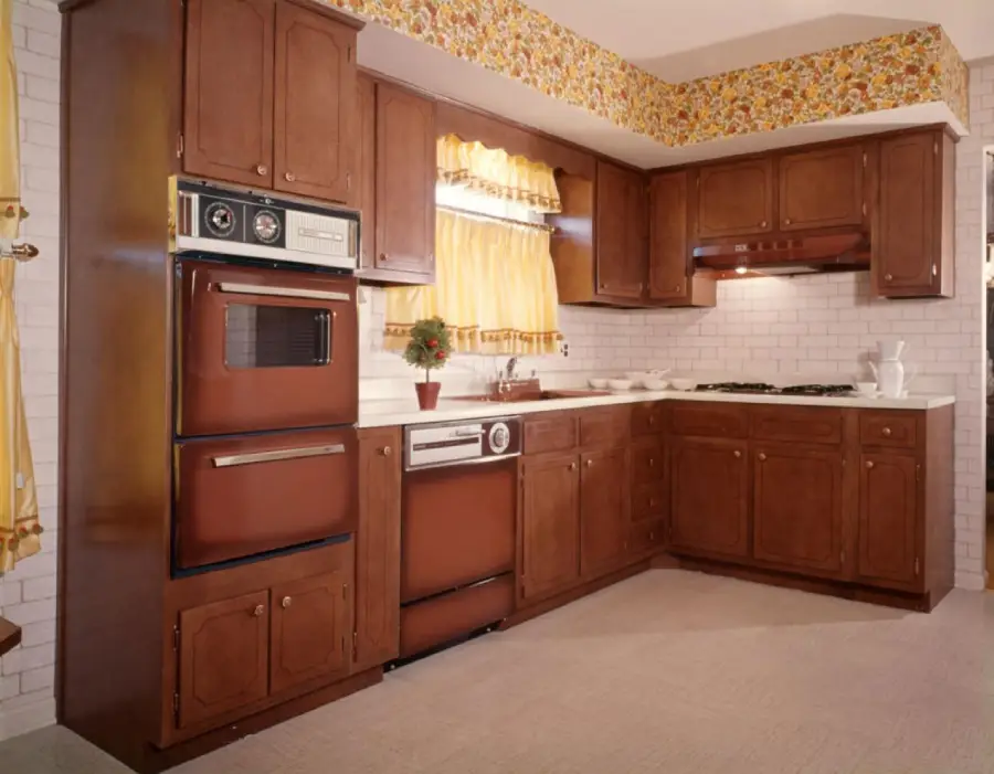 A retro kitchen with brown wood cabinets, vintage appliances, yellow floral wallpaper border, yellow curtains, white brick backsplash, and a small plant on the countertop.