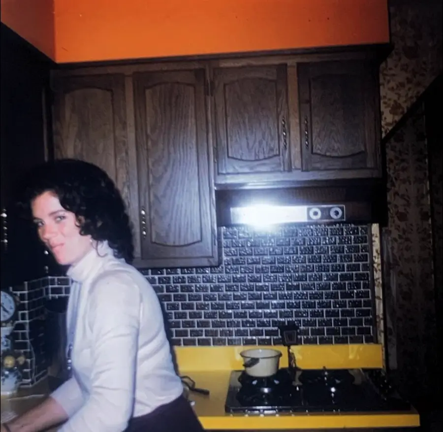 A woman in a white turtleneck stands in a retro kitchen with dark wood cabinets, a yellow countertop, and a stovetop with a pot. The backsplash is black and white tile.