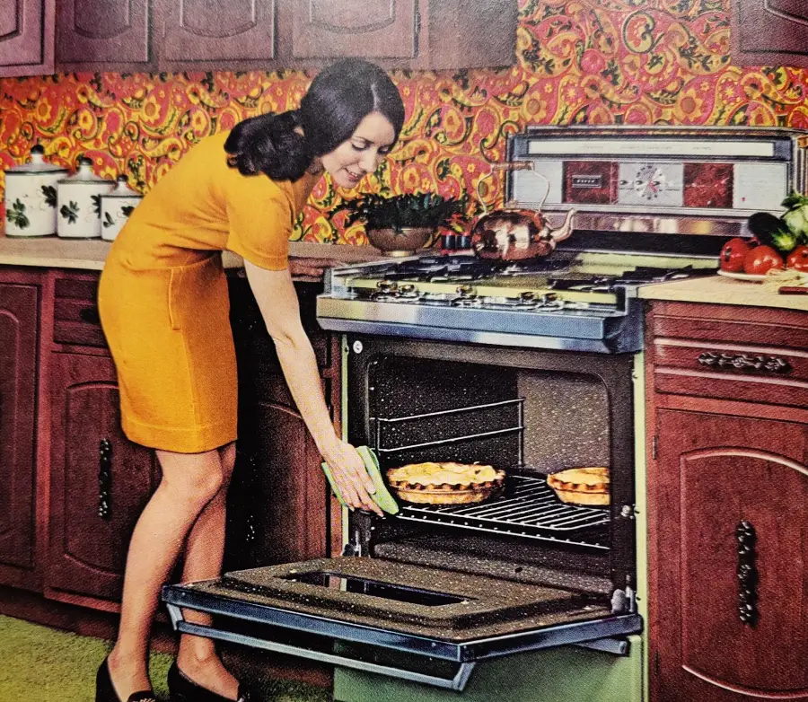 A woman in a yellow dress places a pie in a vintage oven in a retro kitchen with patterned wallpaper, wooden cabinets, and various kitchen items on the countertop.