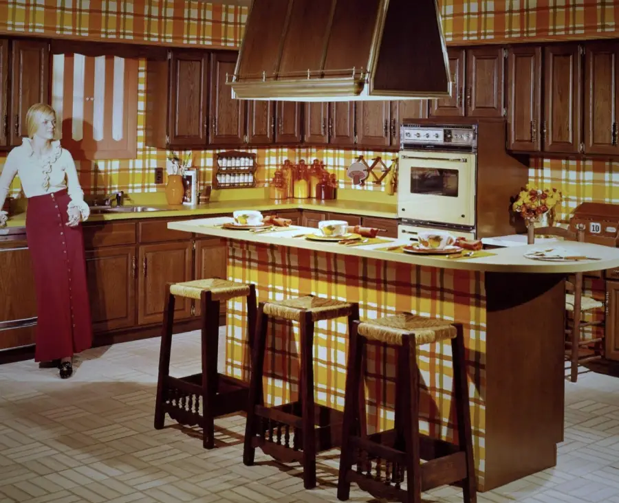 A woman stands in a vintage kitchen with brown cabinets, yellow and orange plaid wallpaper, a central island with three stools, and retro appliances. The décor is bold and patterned, typical of 1970s interior design.