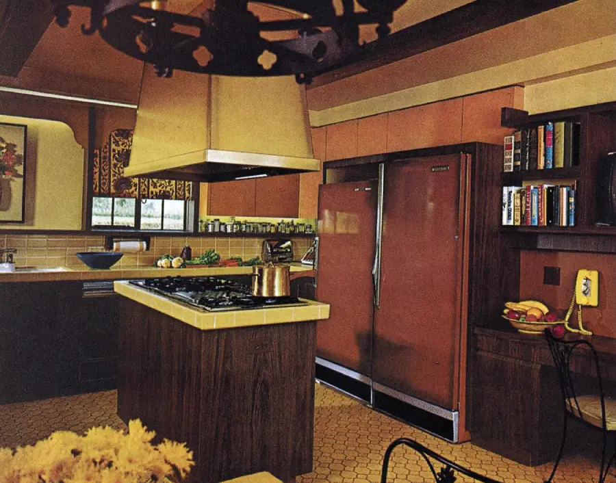 A retro 1970s kitchen features earth tones, brown cabinets, a yellow phone, double brown refrigerator, open shelving, and a kitchen island with a stovetop and copper pot.