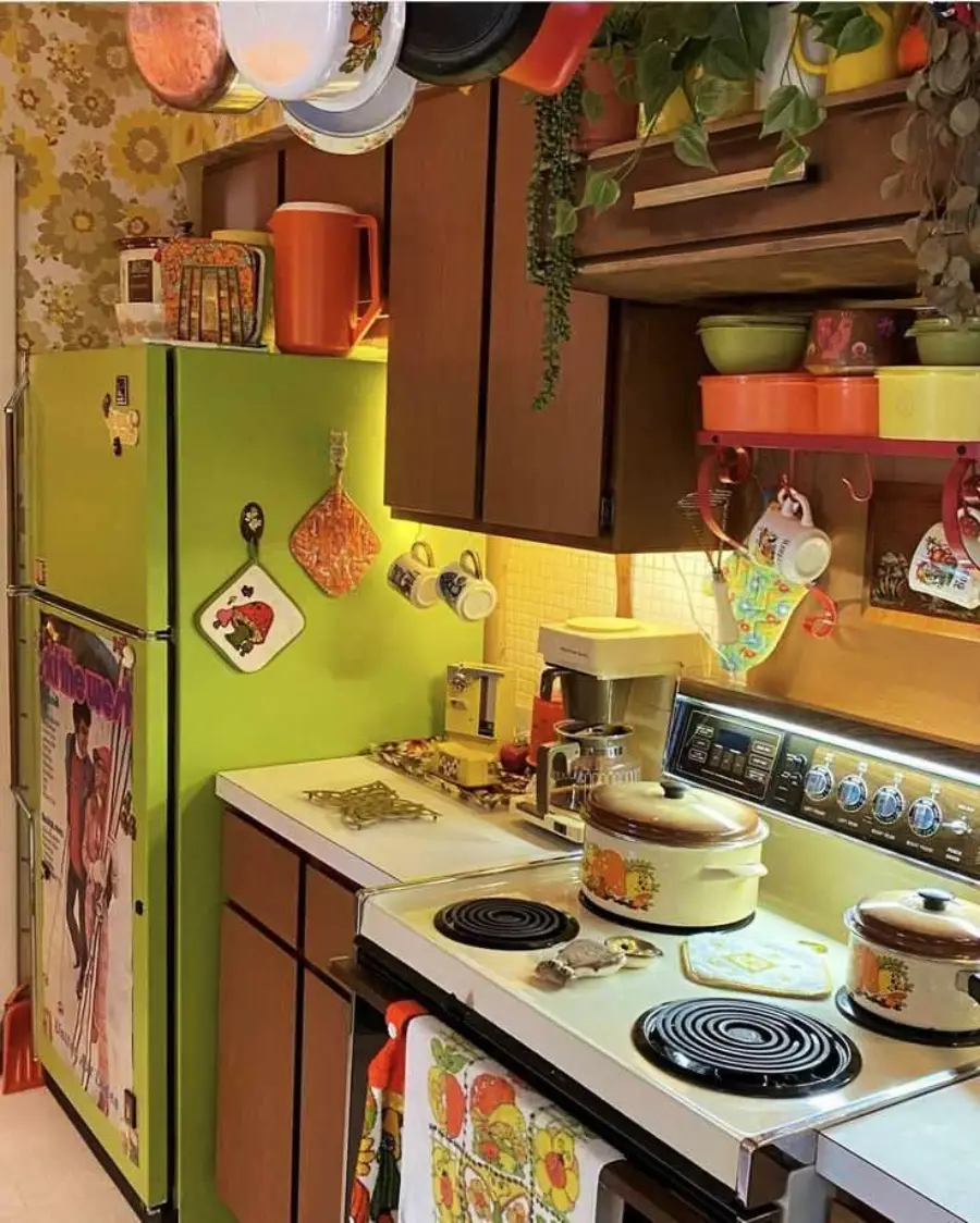 A retro kitchen features green appliances, brown cabinets, floral wallpaper, hanging pots, and colorful utensils. The stove has vintage cookware and the fridge displays magnets and pot holders. The space is cozy and vibrant.