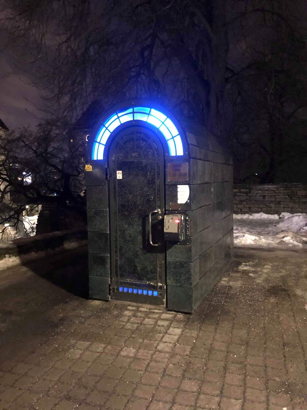 A public restroom with a glowing blue arch above the door stands on a snowy, dimly lit street at night.
