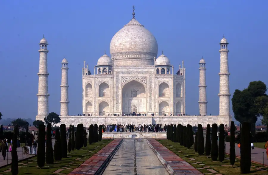 The Taj Mahal, a white marble mausoleum with minarets, stands under a clear blue sky. Reflecting pool and manicured gardens lead up to the entrance, with visitors gathered near the building.