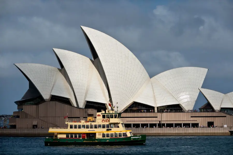 A yellow and green ferry passes in front of the Sydney Opera House, with its iconic white, sail-shaped roof structures, under a cloudy sky.