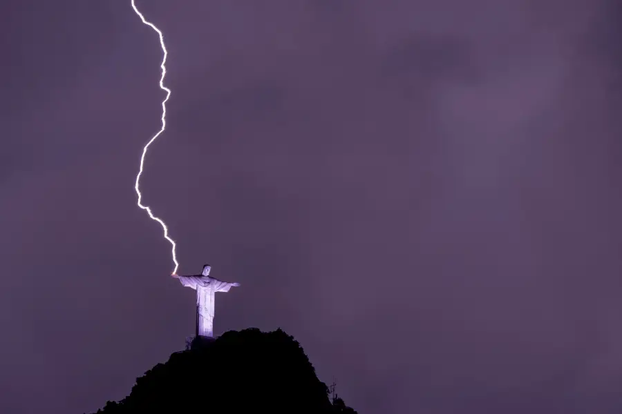 A bolt of lightning strikes the outstretched hand of the Christ the Redeemer statue atop a dark hill, illuminating the night sky with a dramatic purple hue.