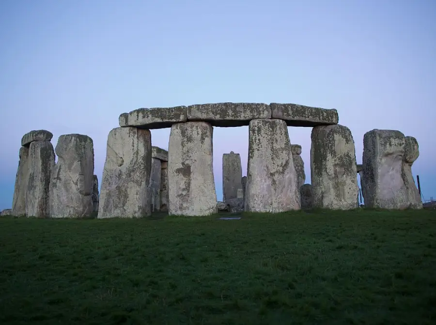 Stonehenge, a prehistoric monument in England, with large upright stones topped by horizontal lintels, stands on a grassy field under a clear blue sky at dusk.