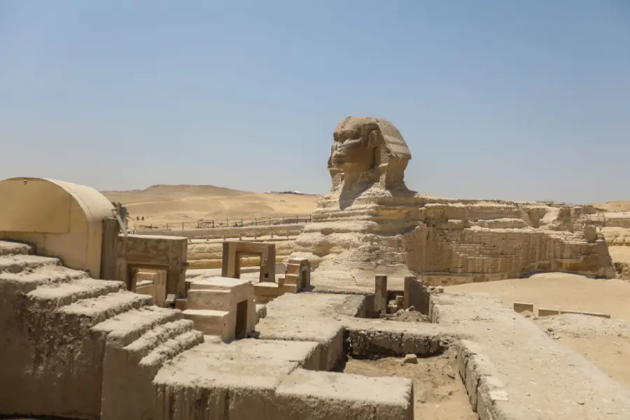 The Great Sphinx of Giza stands in a sandy desert landscape, surrounded by ancient stone ruins and structures under a clear blue sky.
