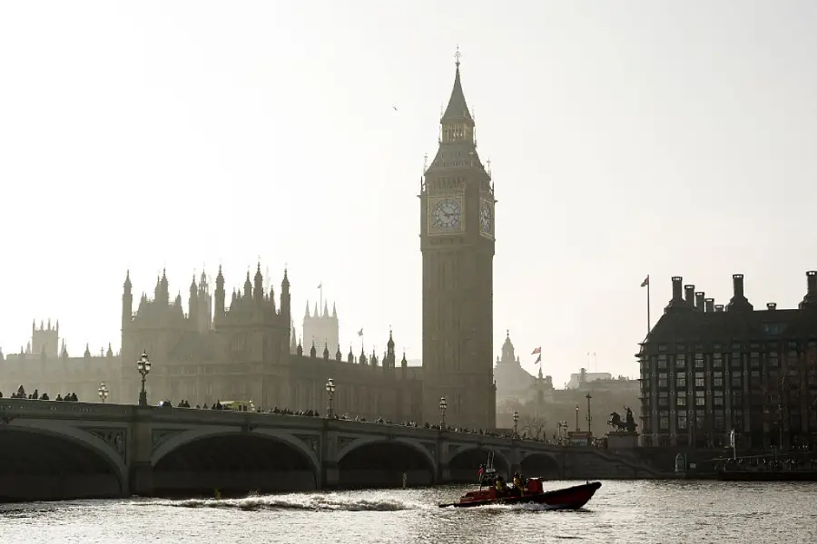 A boat moves along the River Thames in front of the Houses of Parliament and Big Ben in London, with Westminster Bridge and people silhouetted against a hazy sky.