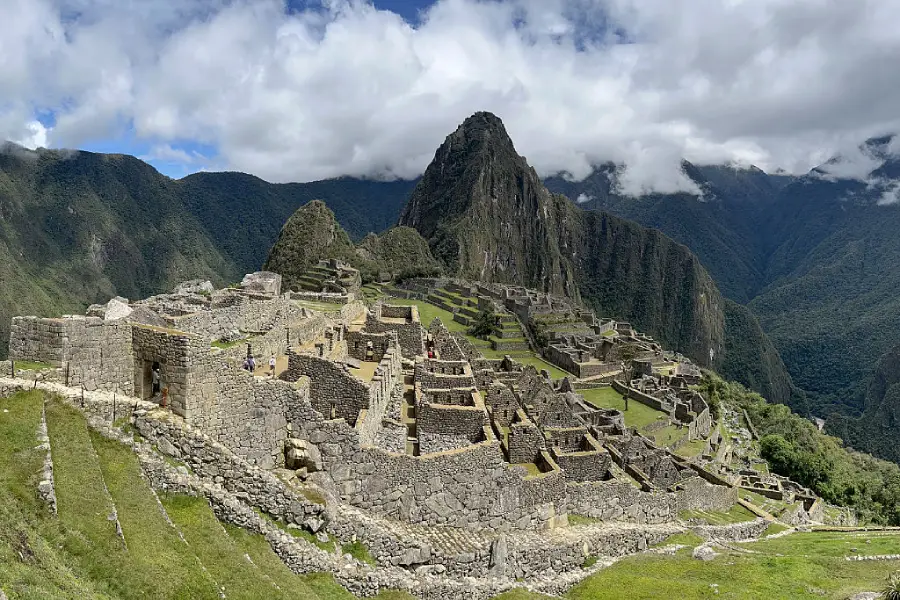 Ancient stone ruins of Machu Picchu sit atop a lush green mountain ridge in Peru, with a dramatic peak and cloudy sky in the background.