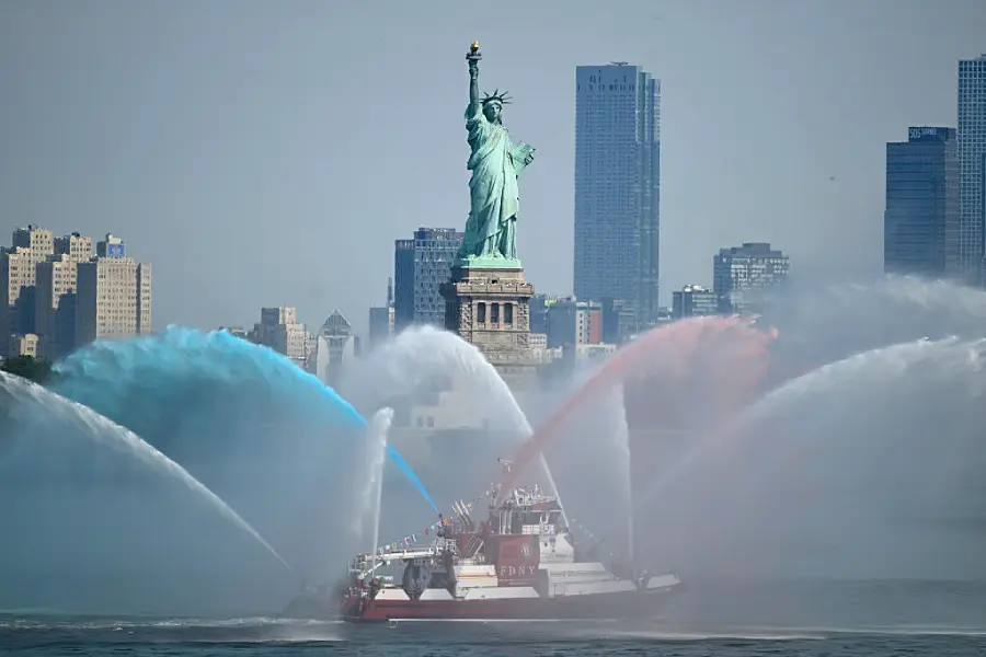 A fireboat sprays red, white, and blue water jets in front of the Statue of Liberty, with skyscrapers and a hazy sky in the background.
