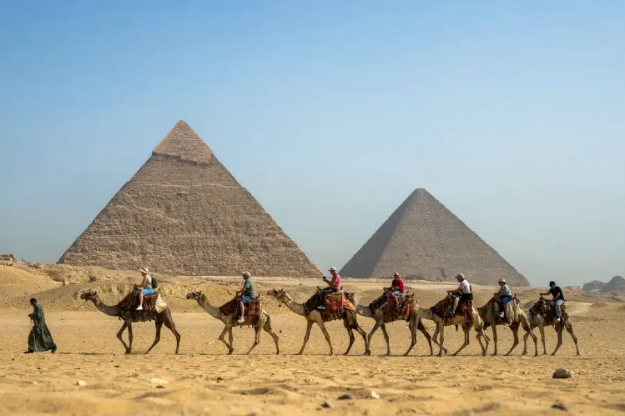 A group of people riding camels passes in front of the pyramids of Giza on a sunny day, with sand and clear blue sky in the background.
