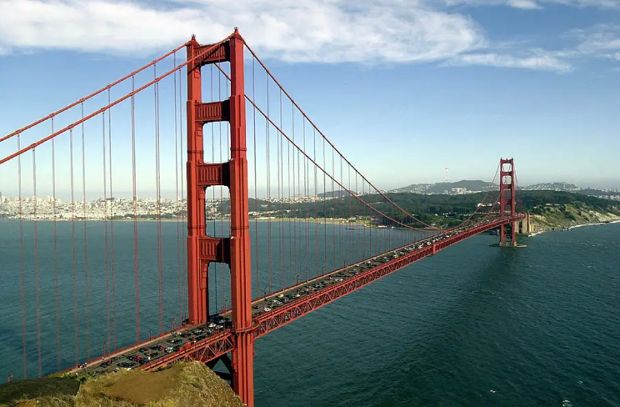 The Golden Gate Bridge, a large red suspension bridge, spans across blue water with a city and hills visible in the background under a partly cloudy sky.