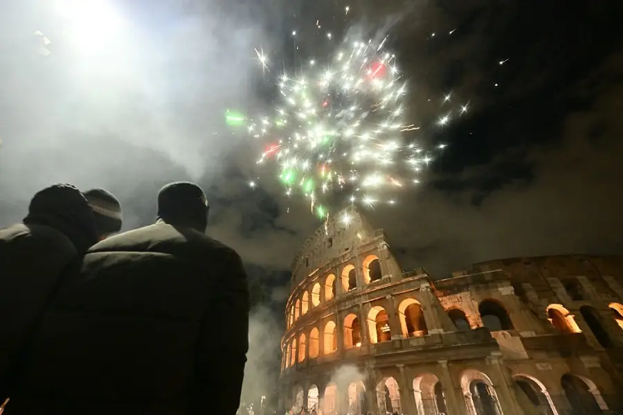 People watch colorful fireworks lighting up the night sky above the illuminated Colosseum in Rome, with clouds and smoke partially visible in the scene.