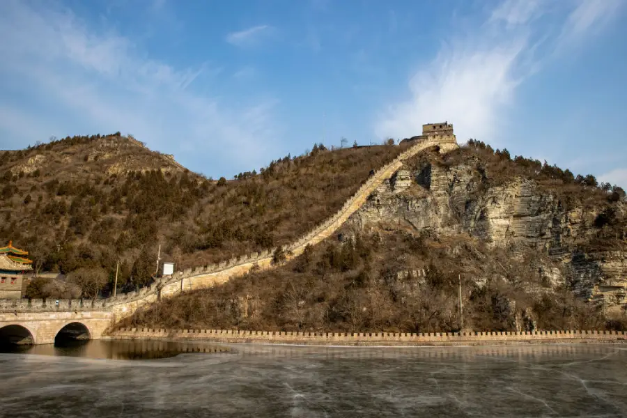 The Great Wall of China stretches over a rocky hilltop, with a watchtower visible, under a blue sky. A stone bridge crosses a frozen river in the foreground.