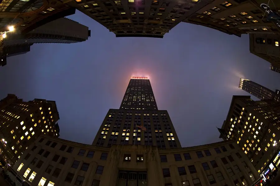 A fisheye view of tall buildings at dusk, with the central skyscraper’s top glowing against a dark, cloudy sky and surrounding structures lit by many windows.