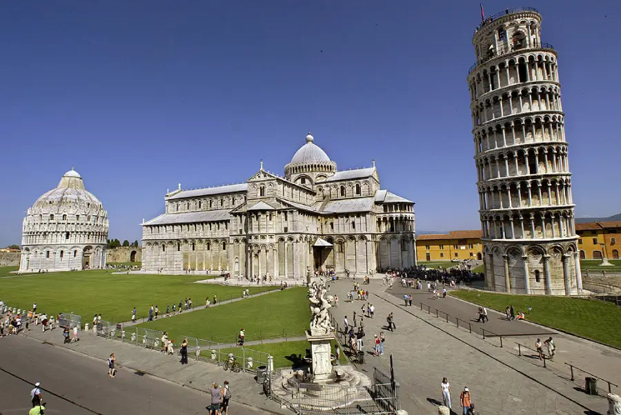 The Leaning Tower of Pisa, Pisa Cathedral, and Baptistery are pictured on a sunny day with blue sky, green grass, and many tourists walking around the historic site in Pisa, Italy.