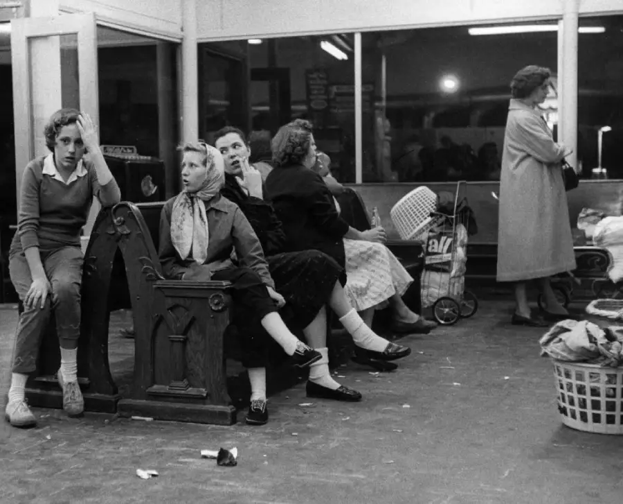Several women sit on benches in a waiting area, some looking tired or anxious. One woman stands by the window in a coat. Laundry baskets and bags are scattered on the floor.