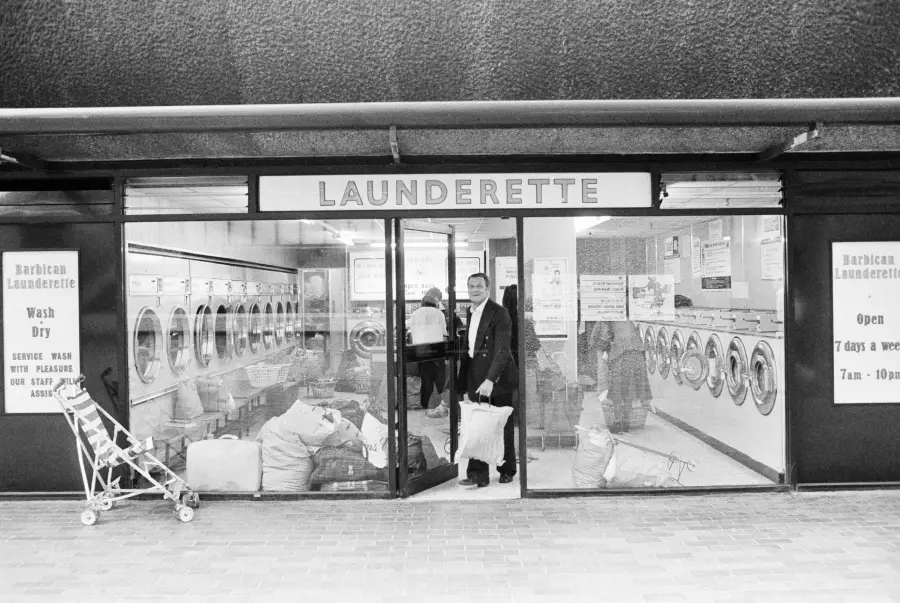 Black and white photo of a launderette with many washing machines, laundry bags on the floor, a man holding clothes, and an empty stroller outside. Signs detail services and opening hours.