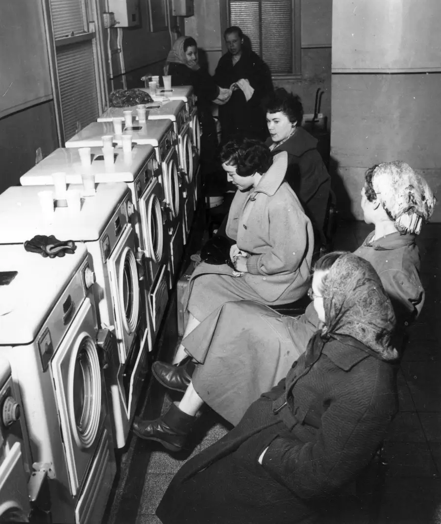Women sit on chairs in front of washing machines in a laundromat, wearing coats and headscarves, waiting for their laundry. Cups are placed on top of the machines, and one man stands in the background.