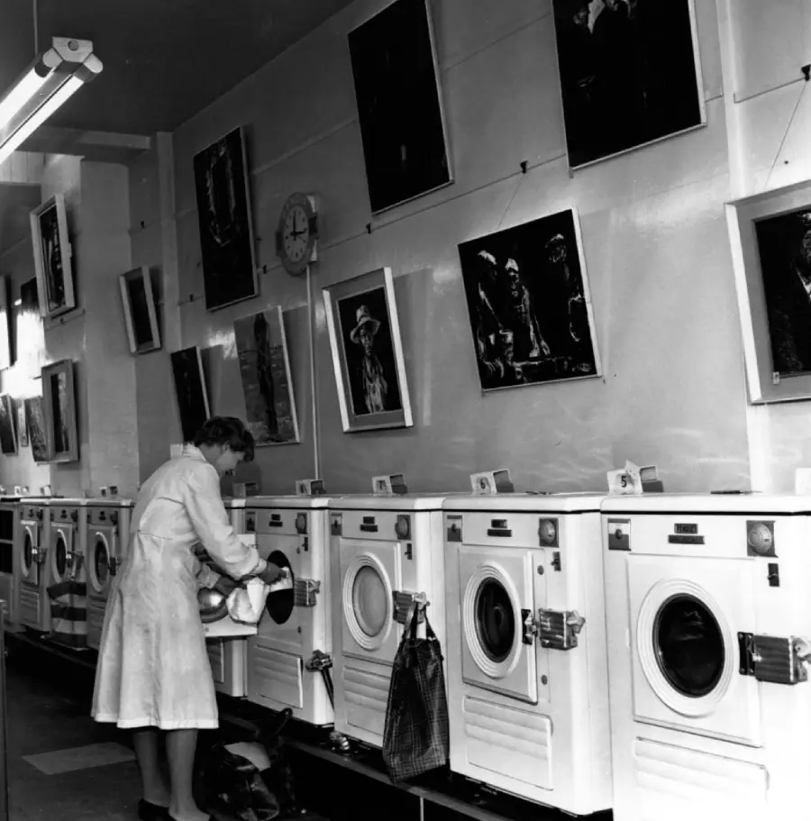 A woman loads laundry into a washing machine in a laundromat. The wall above the machines is decorated with framed photographs. A clock is visible on the wall.
