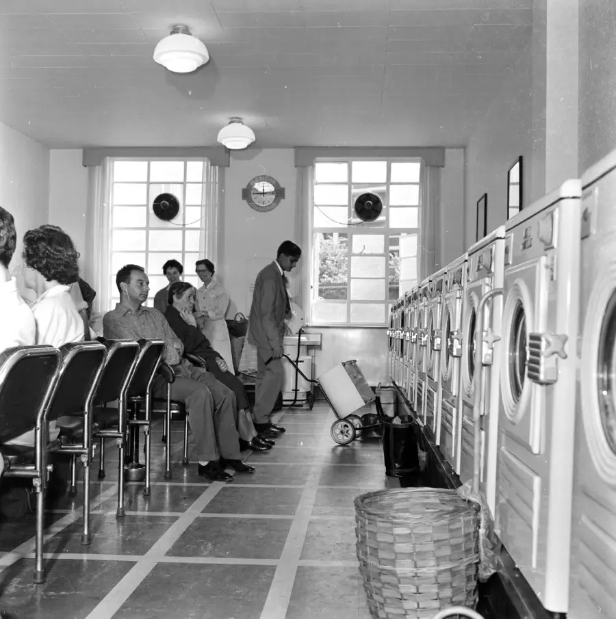 Black-and-white photo of people sitting in chairs at a laundromat, facing a row of washing machines. Some people stand by the machines, while laundry baskets are on the floor. Large windows let in natural light.