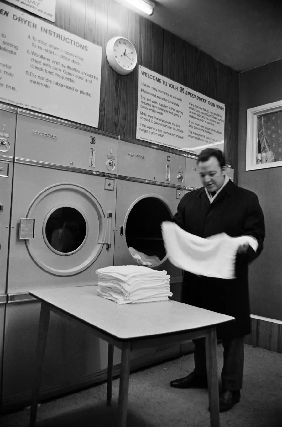 A man in a coat folds laundry at a laundromat, standing by large dryers with a stack of folded towels on a table. Wall signs and a clock are visible above.