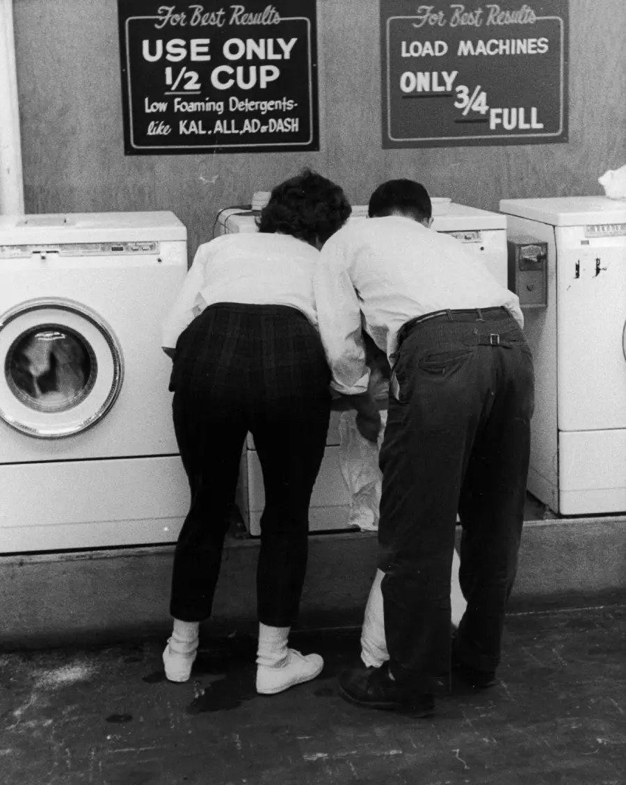A woman and a man lean over a washing machine, possibly loading laundry, in a laundromat. Signs above the machines give detergent and load advice. The setting appears to be from an earlier decade.