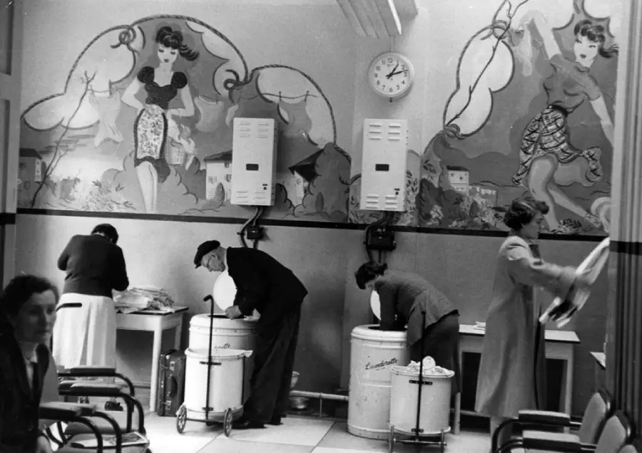 Four people do laundry in a room with washing machines; two large murals of women decorate the wall above, and a clock shows the time.