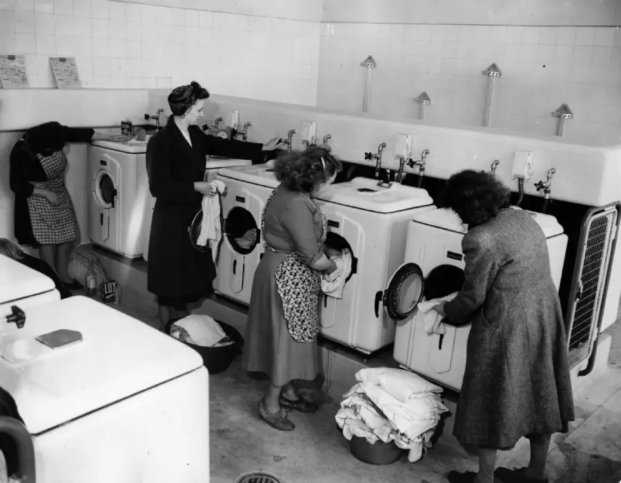 Four women using washing machines in a laundromat, sorting and loading laundry. The setting appears vintage, with tiled walls, baskets of clothes, and old-fashioned washing machines.