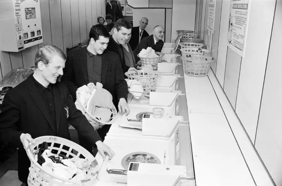 Four men with laundry baskets stand in line using washing machines at a laundromat. Three men in the background watch and smile. The photo is black and white and appears to be from the mid-20th century.