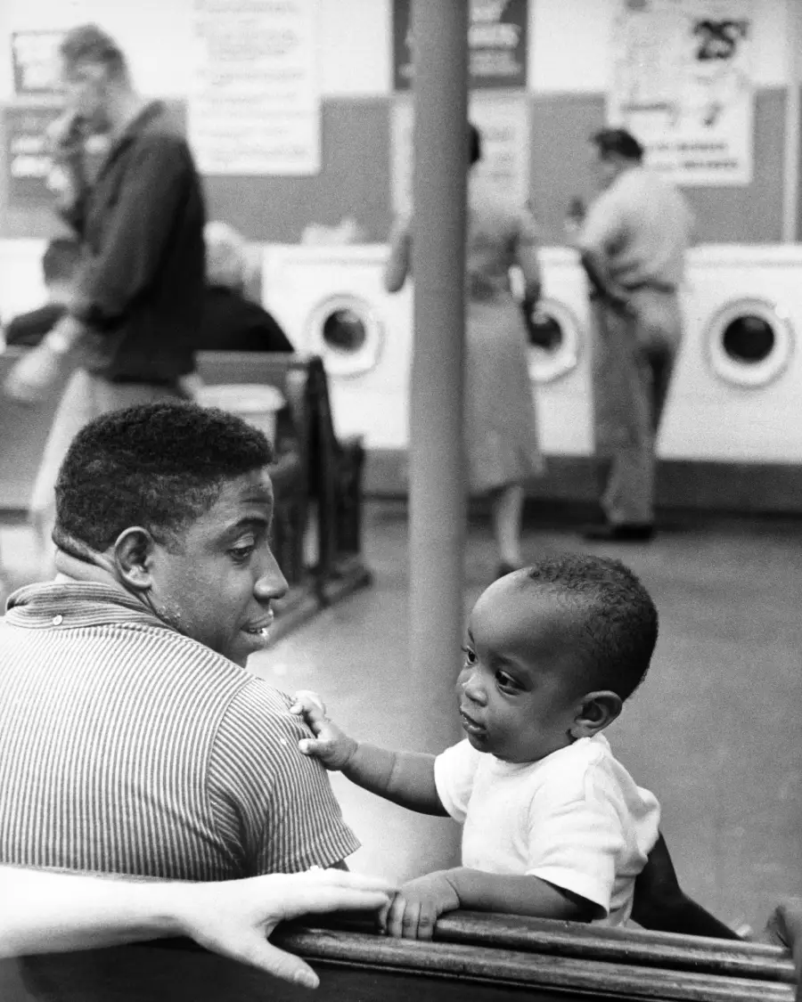 A man and a young child sit on a bench in a laundromat, sharing a moment together. Washing machines and people are blurred in the background.