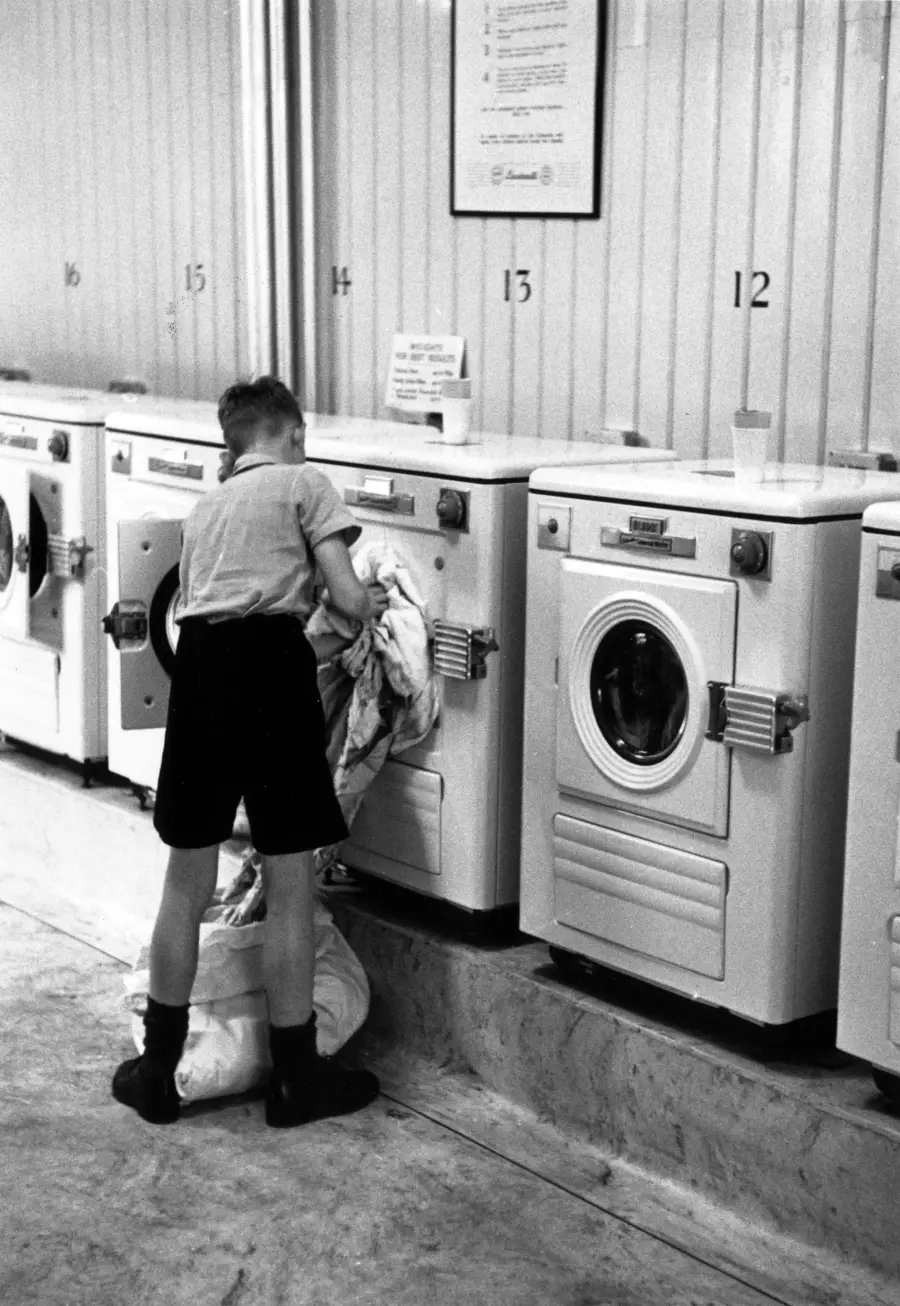 A young boy in shorts loads laundry into a washing machine at a laundromat, with numbered machines lined up along the wall.
